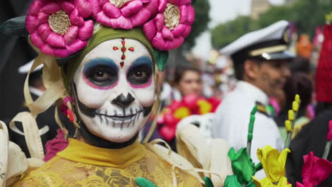 Slow-motion-footage-of-a-woman-in-a-colorful-traditional-outfit-with-sugar-skull-makeup-and-holding-flowers-at-the-Day-of-The-Dead-Parade-in-Mexico-City,-Mexico