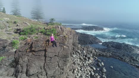 Aerial-drone-flying-forward-pass-woman-sitting-on-rock-cliff-to-Kiama-Rock-Pool-on-a-sunny-misty-morning-with-waves-breaking-on-rocks