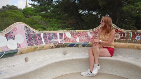 A-modern-woman-wearing-a-smart-top-and-red-shorts,-and-branded-items-like-adidas-shoes-and-a-beautiful-wristwatch-waiting-at-Park-Guell-in-Barcelona-during-the-daytime