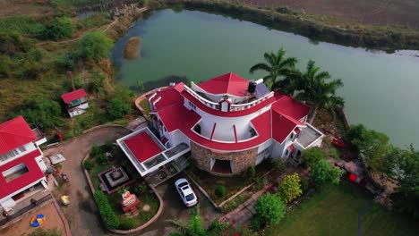 Rotating-aerial-drone-view-of-a-complex-of-summer-houses-for-vacations,-surrounded-by-a-large-bluish-lake,-with-a-parking-lot-and-abundant-green-vegetation-in-Vadodara,-India