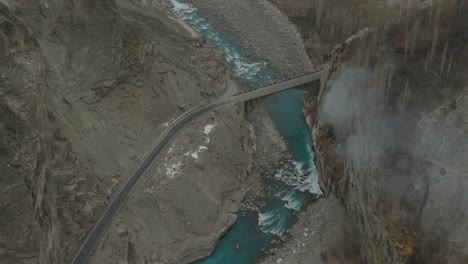 Luftaufnahme-über-Dem-Türkisfarbenen-Fluss-Im-Hunza-Tal-Mit-Rauch,-Der-In-Der-Nähe-Der-Brücke-Aufsteigt