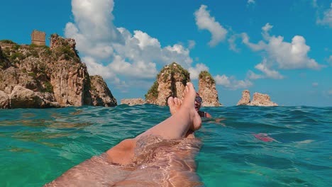 Fpv-of-female-legs-and-feet-relaxing-while-floating-on-sea-water-with-Scopello-Stacks-or-Faraglioni-in-background