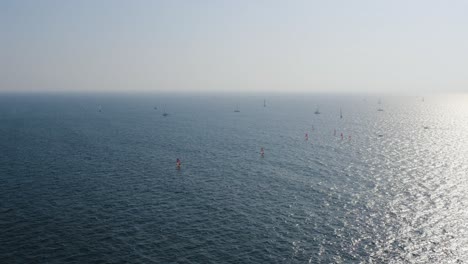 Group-of-windsurfers-with-red-sails-surf-over-calm-and-clear-Mediterranean-sea-as-a-speedboat-passes-at-high-speed-and-the-sunlight-sparkles-on-the-water