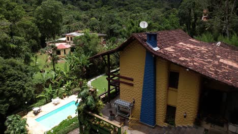 Colorful-resort-aerial-ascend-revealing-lush-tropical-garden-greenery-with-swimming-pool-on-a-mountain-side-with-picturesque-stone-hotel-building-and-tile-roof