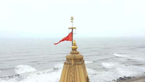 Aerial-backward-moving-shot-of-saffron-colored-Hindu-flag-flying-on-top-of-Somnath-temple-located-in-Prabhas-Patan,-Veraval-in-Gujarat,-India