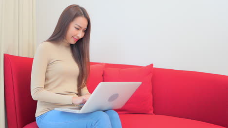 Asian-woman-in-30th-typing-on-laptop-computer-holding-it-on-laps-sitting-on-red-home-sofa
