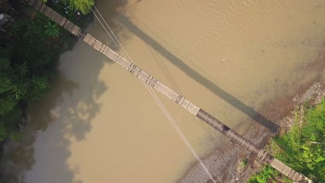 Luftaufnahme-Einer-Hängebrücke-über-Den-Fluss-Mit-Motorradüberquerung-Am-Nebligen-Morgen-Mit-Sonnenstrahl,-Indonesien