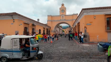 Antigua,-Guatemala--Shot-of-car,-bike-and-auto-passing-by-the-street-with-view-of-Santa-Catalina-Arch-in-the-background-on-a-cloudy-day