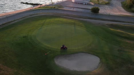 An-aerial-view-of-a-person-riding-a-lawnmower-cutting-the-grass-of-a-golf-course,-next-to-a-beach-during-sunrise