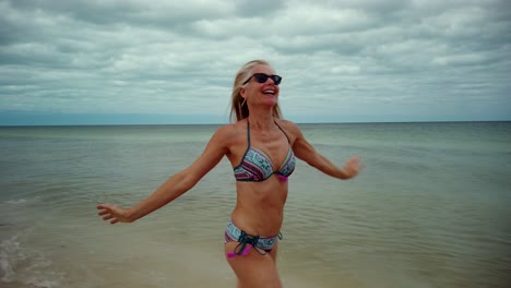 Side-shot-of-playful-blonde-mature-woman-in-sunglasses-and-bikini-running-along-the-shore-laughing-and-putting-her-arms-up-in-the-air-showing-joy