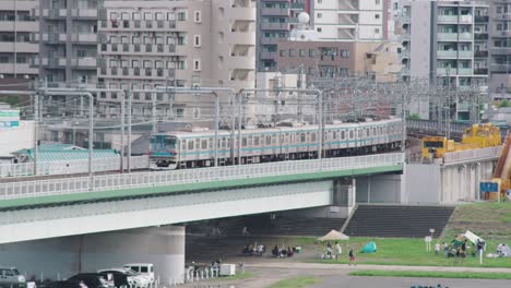 Dos-Trenes-Jr-Pasando-Y-Doblando-Una-Esquina-En-Tamagawa-Con-Gente-Divirtiéndose-En-La-Orilla-Del-Río-En-Tokio,-Japón