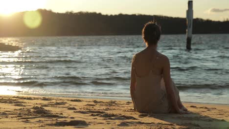 Eine-Junge-Frau-Sitzt-Während-Des-Sonnenuntergangs-In-Einem-Langen-Kleid-Am-Wasser-Und-Spielt-Mit-Dem-Sand