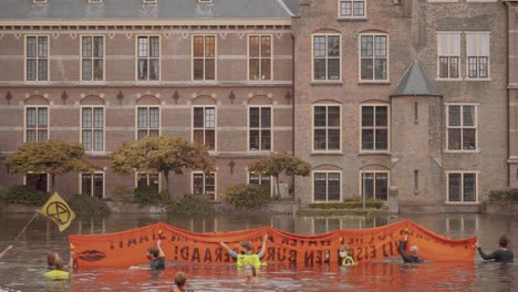Protesters-Holding-Orange-Banner-against-climate-change-swimming-In-Hofvijver-pond,-Water-Facing-Dutch-parliament,-tweede-kamer-at-binnenhof-Building---The-Hague,-Netherlands---slow-motion