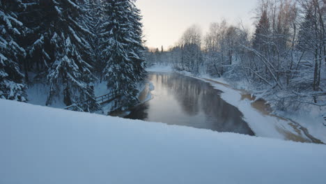 Revealing-motion-shot-of-river-by-snowy-forest-in-wintertime-Finland