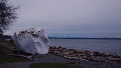 Pan-on-a-giant-white-boulder-on-white-rock-beach-covered-with-driftwood-overlooking-the-ocean-and-mountains-in-the-distance-during-evening-clouds-trees-dark-horizon