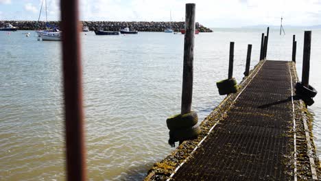 Relaxing-wooden-harbour-jetty-walkway-tranquil-scene-over-blue-sky-calm-sea-tide-right-dolly-passing-fence-bars