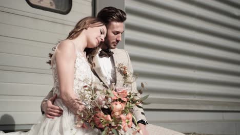 Wedding-couple-sitting-thinkful-in-front-of-a-grey-warehouse-gate-cuddling-enjoying-the-moment-and-kissing