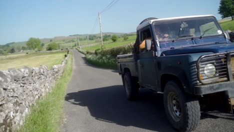 Panning-slow-mo-shot-of-old-blue-Land-Rover-defender-driving-past-on-a-typical-British-countryside-road