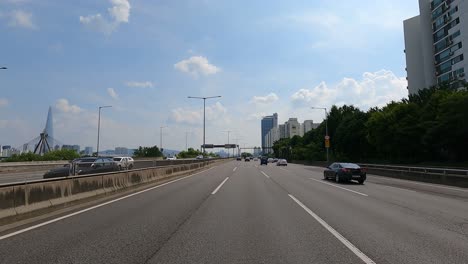 Driver's-POV-Road-traffic-along-Gangbyeonbuk-ro-road-near-Han-river,-Lotte-tower-and-Olimpic-Bridge-on-background,-Seoul,-Korea,-Jun-27,-2021-afternoon-summer-day