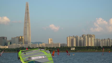 Windsurfers-surfing-on-the-Han-River,-Lotter-World-Tower-on-the-background-at-sunset,-Jamsil,-Seoul,-South-Korea