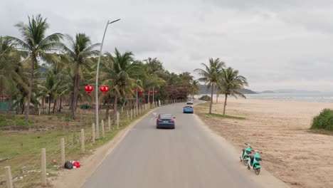 Tesla-Model-3-driving-on-tropical-road-by-the-beach-with-numerous-camper-vans-parked-on-the-roadside