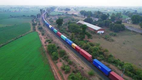 Güterzug-Mit-Blauen,-Gelben,-Roten-Und-Hellblauen-Waggons,-Der-An-Einem-Bewölkten-Tag-Durch-Die-Grünen-Erntefelder-In-Indien-In-Der-Nähe-Einer-Autobahn-Fährt
