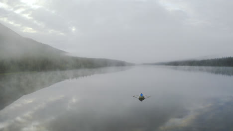Verträumte-Landschaft-Mit-Nebel-über-Dem-Wasser-Und-Einer-Person,-Die-Zwischen-Bergen-Ein-Boot-Rudert