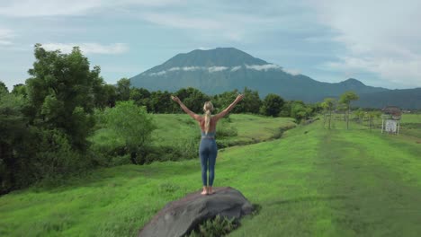 Adventure-woman-running-barefoot-on-grass-field-steps-on-rock-boulder-celebrating-with-arms-up,-breathtaking-view-of-grand-volcano-Agung