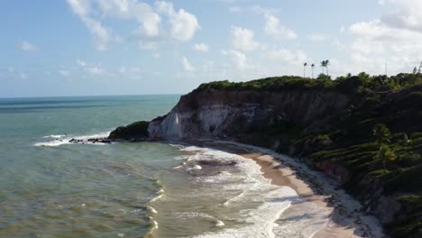 Dolly-out-aerial-drone-shot-of-the-beautiful-tropical-coast-of-Paraiba,-Brazil-near-Joao-Pessoa-on-the-Gramame-beach-point-with-colorful-clay-cliffs,-palm-trees,-and-small-waves-on-a-warm-summer-day