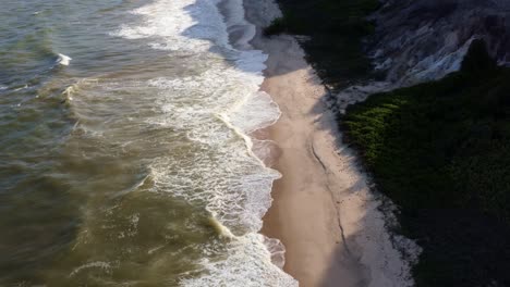 Dolly-in-aerial-drone-top-view-shot-of-the-beautiful-tropical-coast-of-Paraiba,-Brazil-near-Joao-Pessoa-with-small-waves-hitting-the-beach-leading-to-green-bushes-and-a-cliff-on-a-warm-summer-day