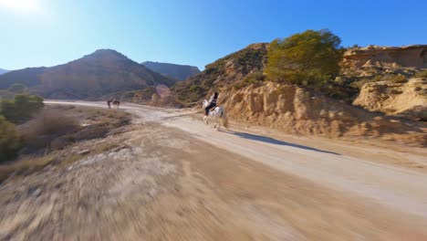 FPV-drone-following-a-woman-riding-a-horse-along-a-country-trail-in-a-sunny,-desert-landscape