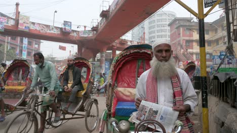 A-man,-a-rickshaw-puller,-reads-a-newspaper-outdoors-as-commuters-drive-and-walk-past-through-the-frame-in-a-busy-street