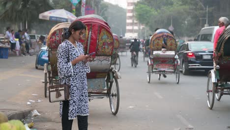 A-woman-uses-a-smartphone-on-the-side-of-the-road-as-vehicles,-including-rickshaws,-drive-past-her-on-a-busy-road-in-Dhaka,-Bangladesh