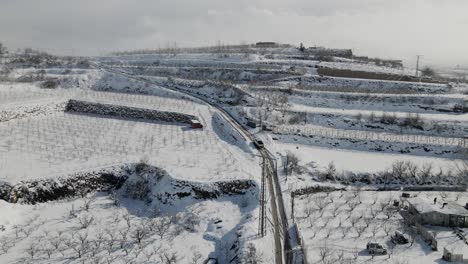Static-aerial-view-of-trucks-driving-up-road-in-snow-of-terraced-vineyard-and-village,-Israel