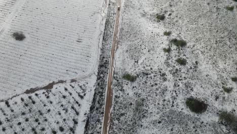 Drone-top-view-following-vehicle-on-dirt-road-after-snow-storm-and-vineyards,-Israel
