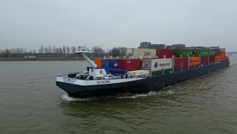 Aerial-Tracking-Shot-Off-Port-Side-Of-Scaldis-Container-Ship-Travelling-Along-Oude-Maas-On-Overcast-Day-In-Dordrecht