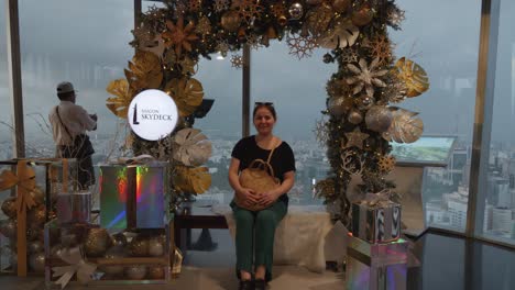 A-woman-in-her-thirties-sits-on-a-bench-surrounded-by-a-festive-Christmas-display-at-the-Skydeck-of-the-Bitexco-Financial-Tower-in-Ho-Chi-Minh,-Vietnam