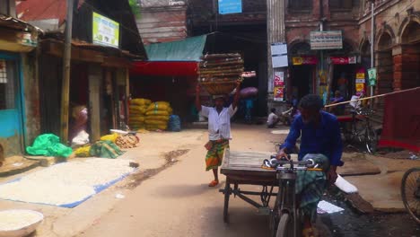 An-Asian-day-laborer-carries-a-sack-or-basket-on-their-head-through-a-busy-market,-showcasing-the-daily-hustle-and-bustle-of-local-workers