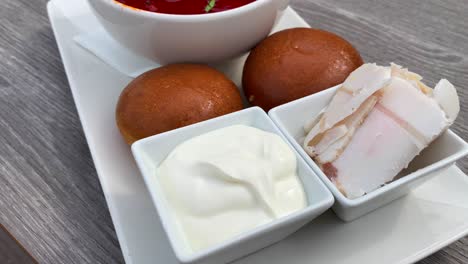 Traditional-Ukrainian-borscht-soup-with-bread-pampushki-buns,-lard-salo-cured-pork-fat-and-sour-cream,-soup-with-beef-and-beetroot,-4K-shot