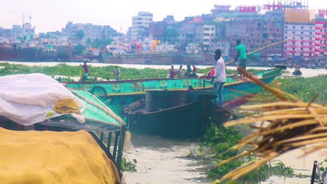 Un-Ajetreado-Río-Buriganga-Fluye-Con-Barcos,-Incluido-Un-Barco-Con-Motor-De-Arrastre,-Con-La-Ciudad-De-Dhaka-Visible-Al-Fondo