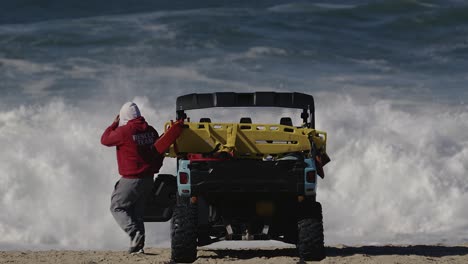 Llifeguard-in-a-red-hoodie-gets-out-of-a-duny-buggy-on-the-beach-in-front-of-huge-waves-in-the-background