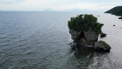 Aerial-view-of-a-stunning-giant,-tree-covered-rock-in-the-ocean-resembling-the-face-of-Jesus-with-waves-crashing-into-it