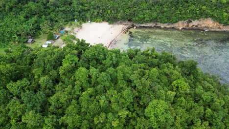 Cinematic-aerial-view-of-tree-covered-hill-revealing-hidden-white-sand-beach-resort-with-stunning-clear-waters