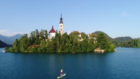 Bled-Island-with-the-Church-and-People-Paddling-at-the-Blue-Lake,-Slovenia-Tiny-Natural-Paradise,-Aerial-View