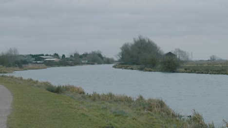 Panning-shot-on-gray-overcast-day-in-the-middle-of-English-winter-of-a-river-with-a-path-and-a-jogger,-Ely-England