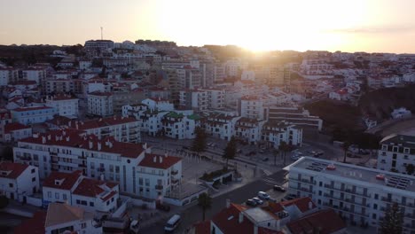 Drone-rising-over-Ericeira-village-with-Sun-flare-through-the-rooftops,-Portugal