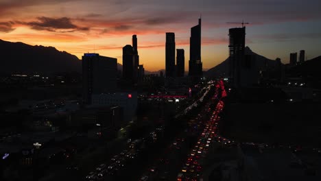 Aerial-view-over-traffic,-towards-silhouette-skyscrapers-in-San-Pedro-Garza-Garcia,-Monterrey,-colorful-dusk-in-Mexico