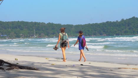 two-barefoot-female-tourists-walk-along-windy-tropical-beach-with-backpacks