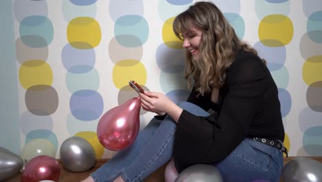 Young-Woman-Sitting-Around-Balloons-and-Holding-a-Phone-in-Hand-Against-Colorful-Background