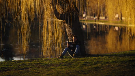 Young-man-reading-a-book,-sitting-under-a-golden-tree-by-a-public-city-park-lake,-at-dusk